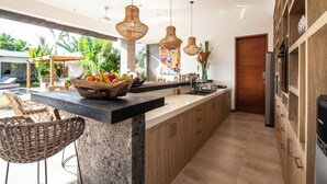 Una cocina moderna con encimera de granito, armarios de madera y una isla grande con frutero. Hay sillas de mimbre y vistas al exterior.