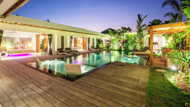 Un área junto a la piscina con terraza de madera, sillas de descanso y un espacio interior bien iluminado.