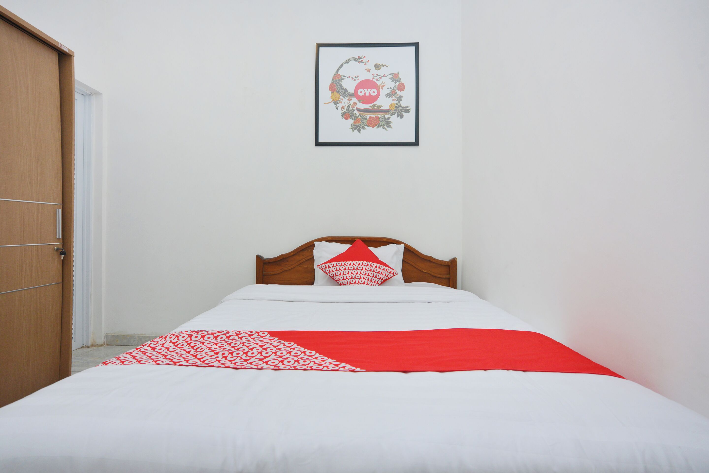 Een net opgemaakt bed met een rood-wit beddegoed, een houten hoofdbord en een ingelijst foto aan de muur.