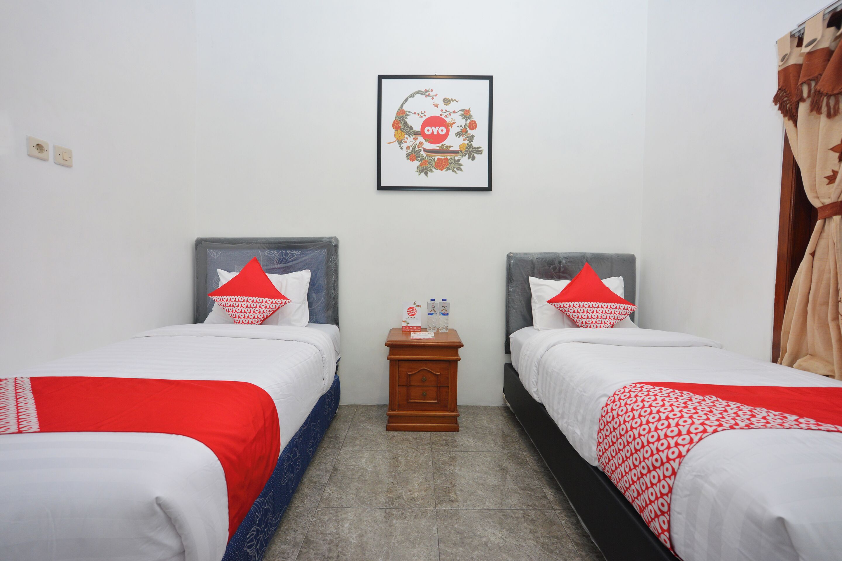 A hotel room with two beds, each with red and white bedding, a small wooden nightstand with a framed picture above, and a curtain on the right side.