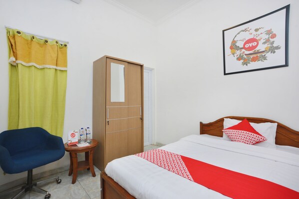 A hotel room with a bed, a wooden wardrobe, a blue chair, a small round table with a bottle and a menu, and a framed picture on the wall.