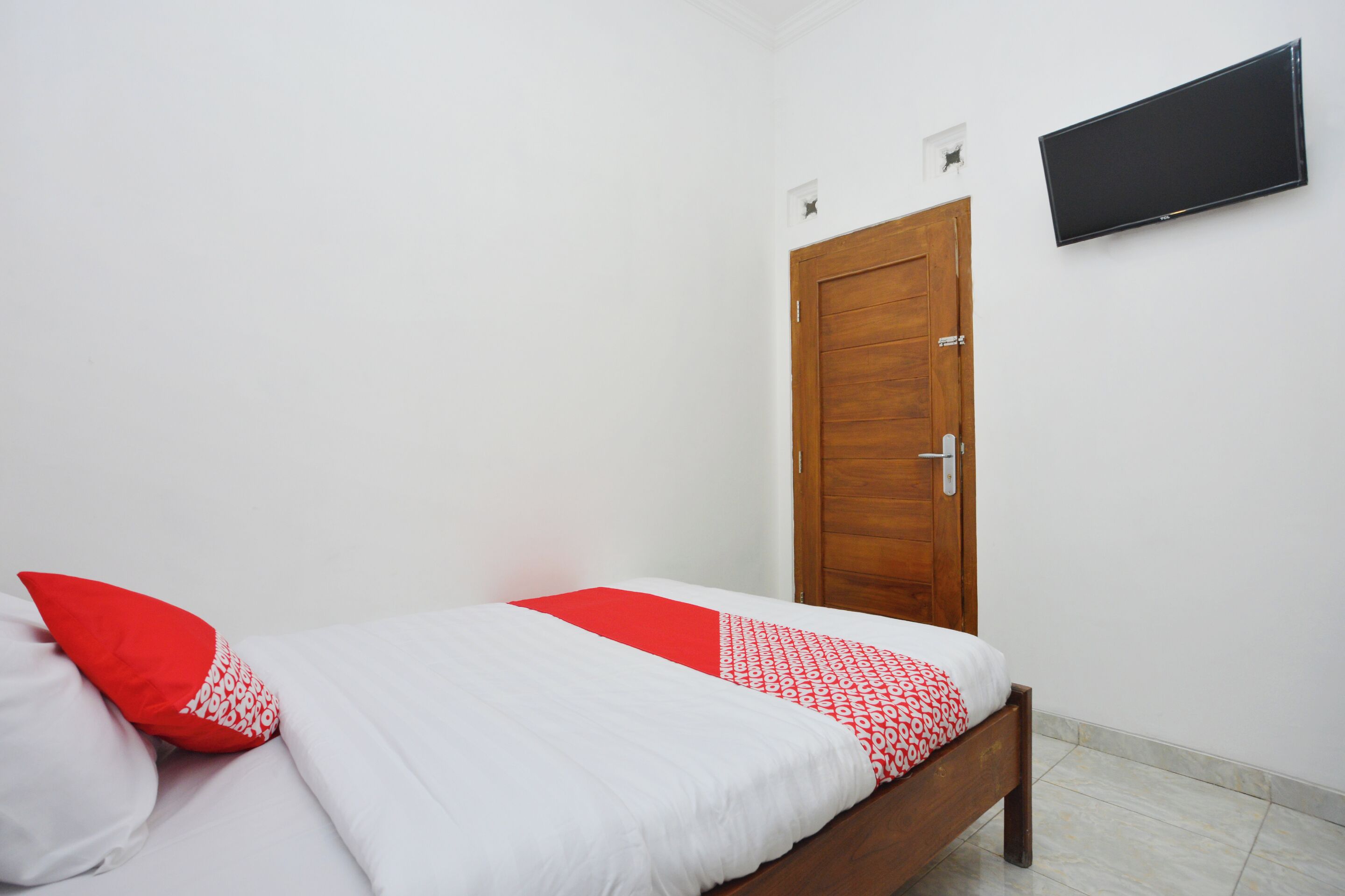 A single bed with white and red bedding, a wooden door, and a flat-screen TV mounted on the wall.