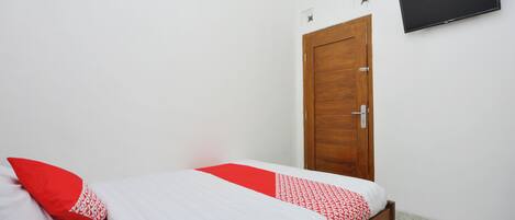 A single bed with white and red bedding, a wooden door, and a flat-screen TV mounted on the wall.