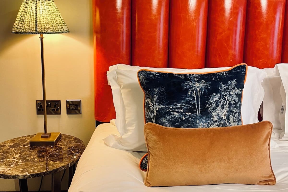 A hotel room with a bed, a lamp, and a wall with a red headboard.