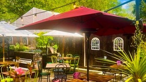 Une terrasse extérieure avec des tables et des chaises en bois, un parasol rouge et des guirlandes lumineuses.