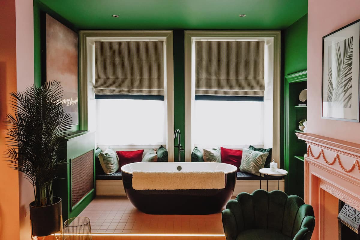 A bathroom with a freestanding bathtub, two windows with blinds, a green chair, and a fireplace.