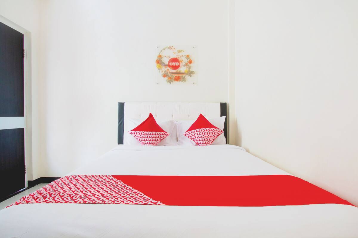 Habitación de hotel con cama blanca, colcha estampada en rojo y blanco y dos almohadas rojas.