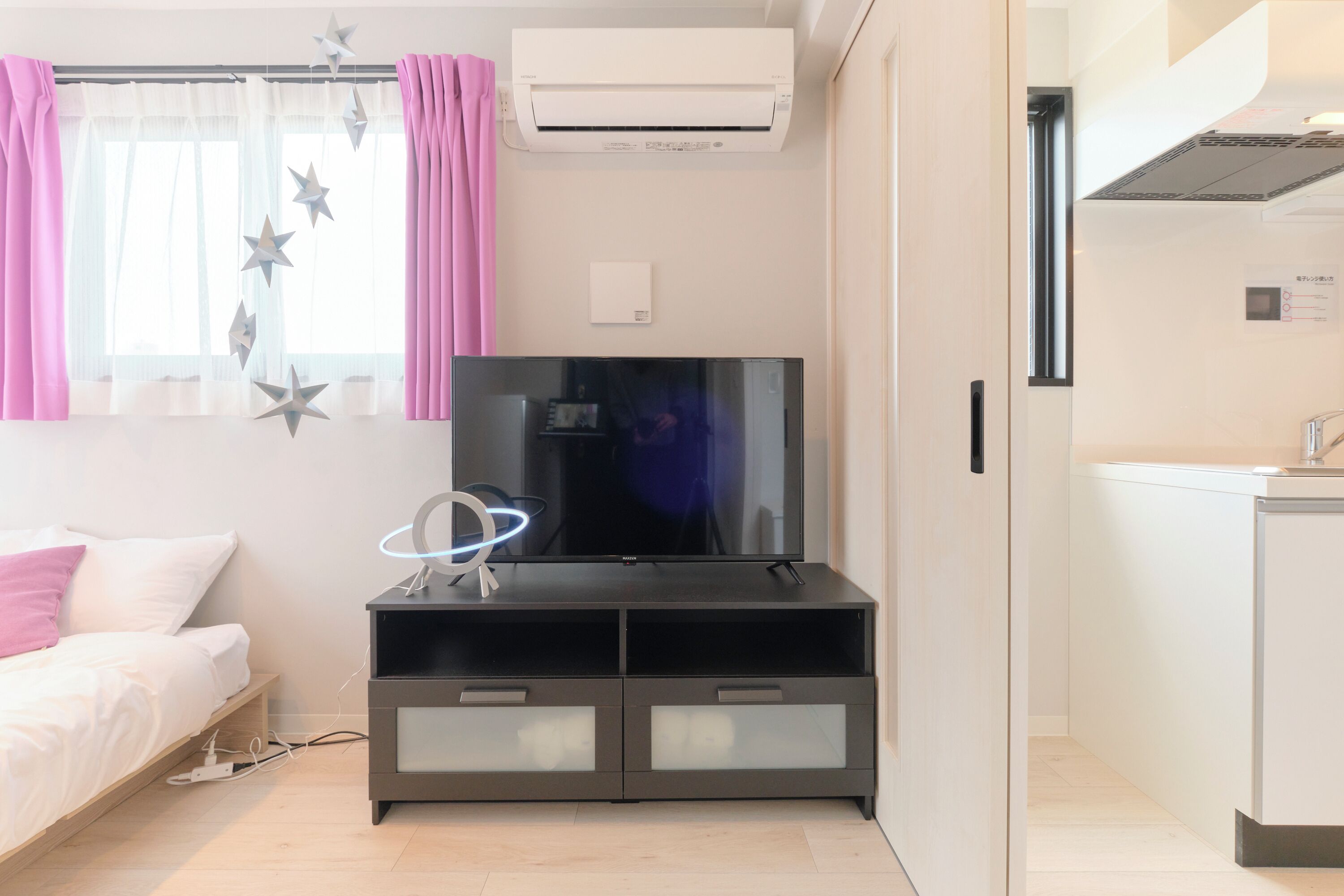 Una habitación de hotel moderna con una televisión de pantalla plana sobre un mueble de madera oscura, una cama con ropa blanca, un escritorio pequeño con una lámpara y una zona de kitchenette.