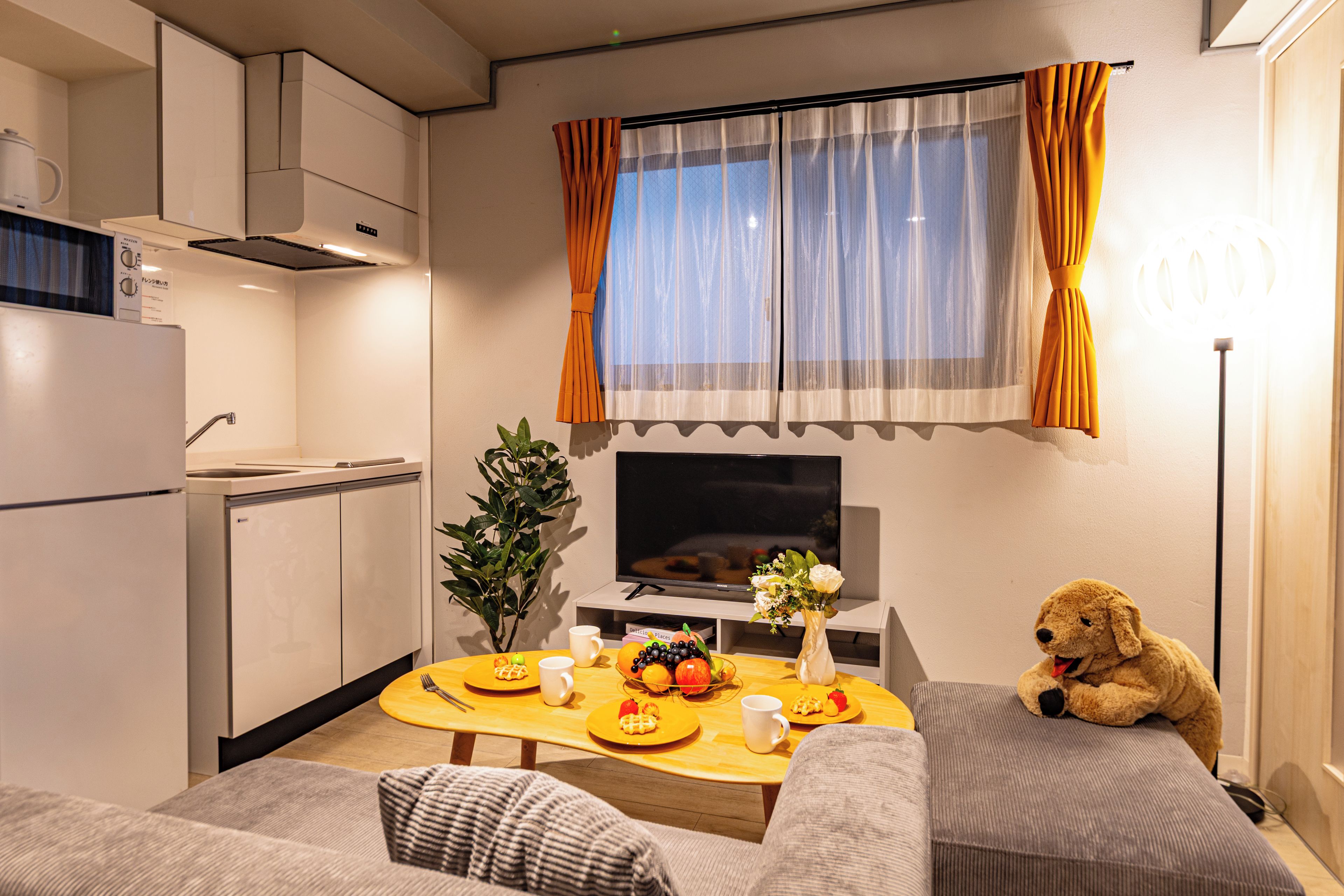 Espaço compacto com cozinha americana, área de jantar com mesa posta para dois, sofá, televisão e um urso de pelúcia.