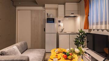 A compact living space with a sofa, a dining table with fruit and a vase, a kitchen area with appliances, and a TV.