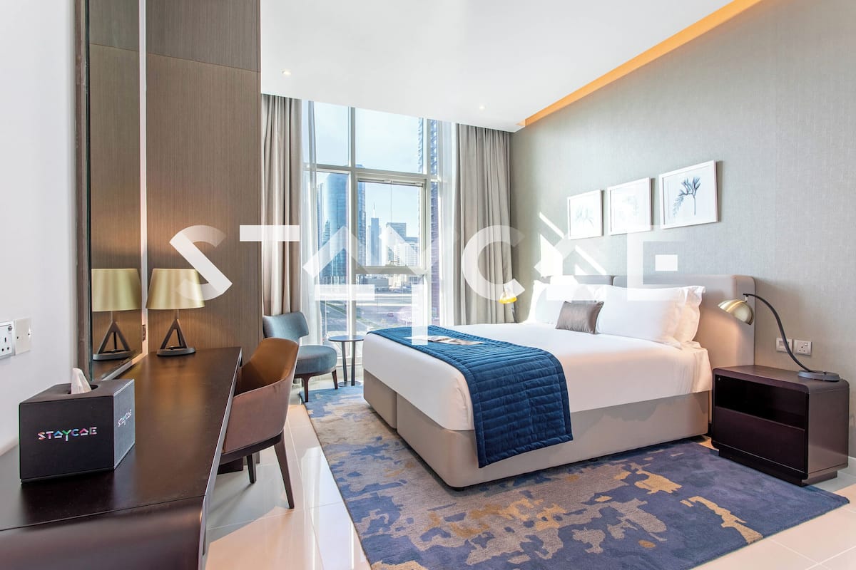 A modern hotel room with a large bed, a desk, a chair, and a view of the city through large windows.