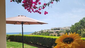 Eine Terrasse mit zwei Liegestühlen und einem Tisch unter einem großen Sonnenschirm, mit Blick auf eine Küstenstadt, die mit Gebäuden und Grünflächen gesäumt ist.