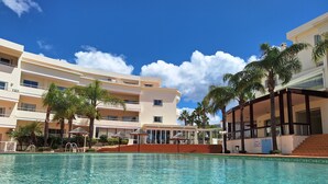 Ein Swimmingpool mit klarem Wasser, umgeben von Palmen und einem mehrstöckigen Hotelgebäude mit Balkonen.