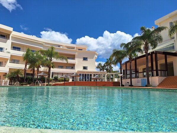 A swimming pool with clear water, surrounded by palm trees and a multi-story hotel building with balconies.