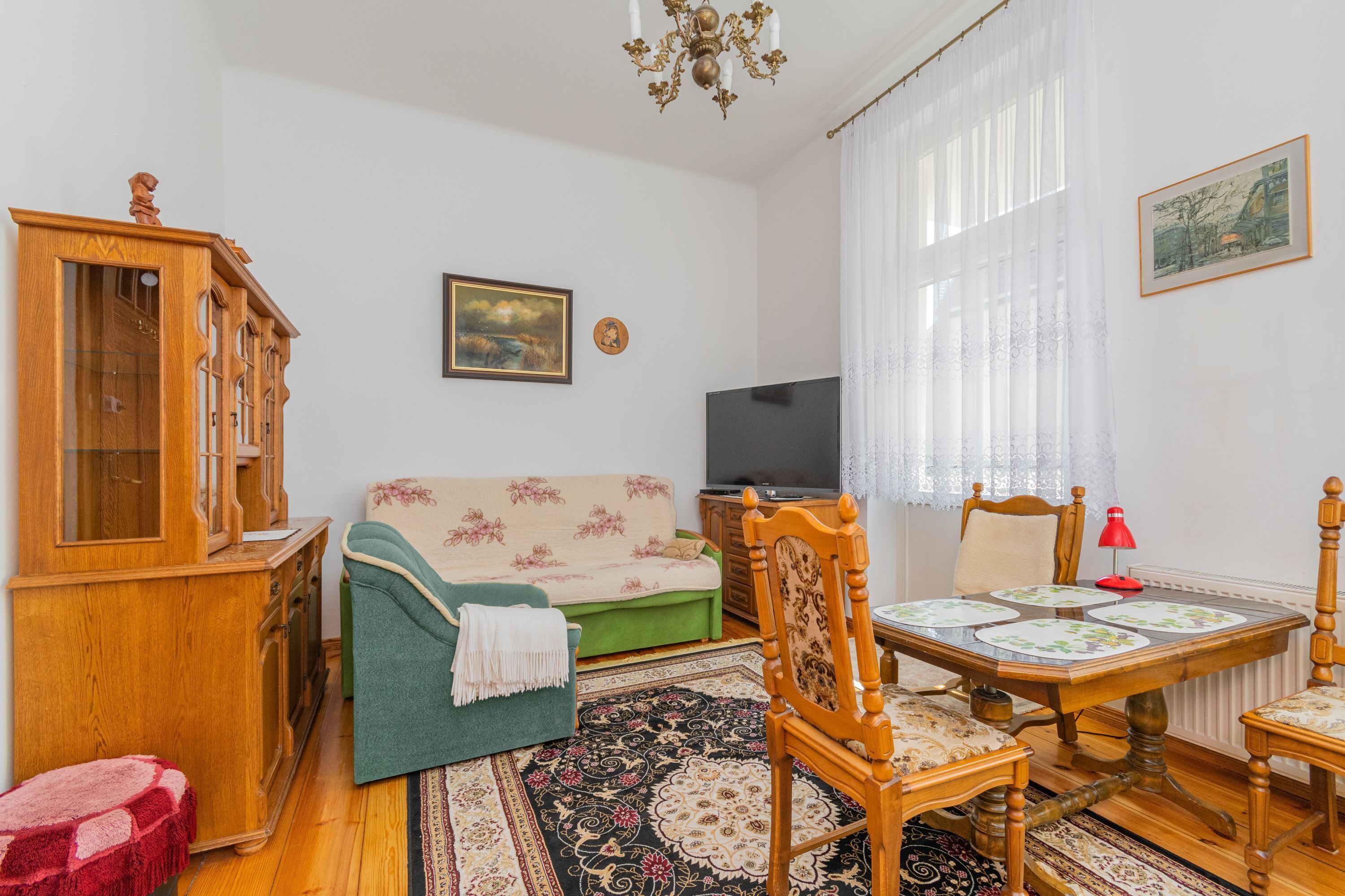 A living room with a wooden cabinet, a sofa, a television, a dining table with chairs, and a patterned rug.