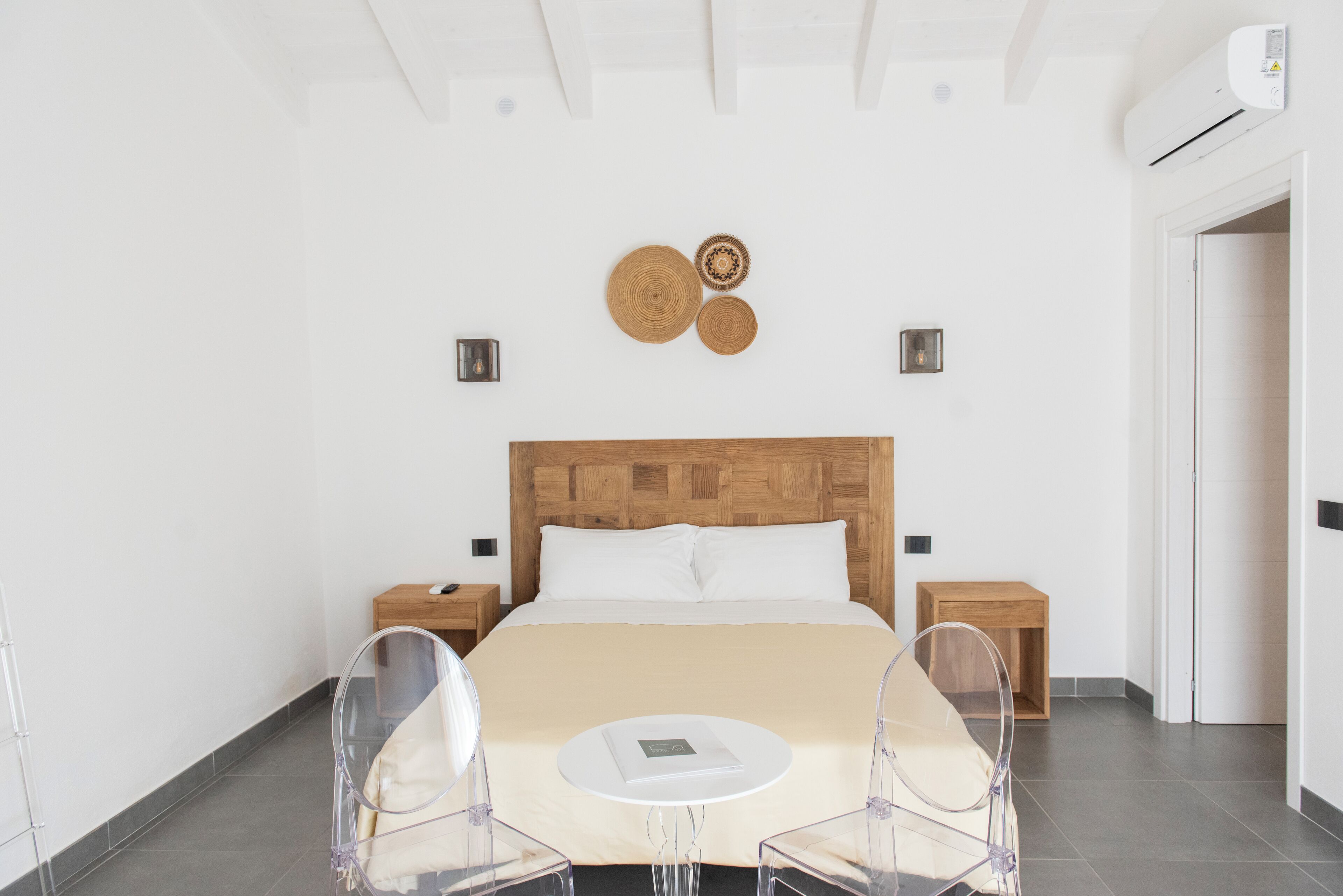 Ein modernes Hotelzimmer mit einem Holzkopfteil, einem Bett mit weißen Laken, zwei Nachttischen und einem klaren Kunststoffstuhl.