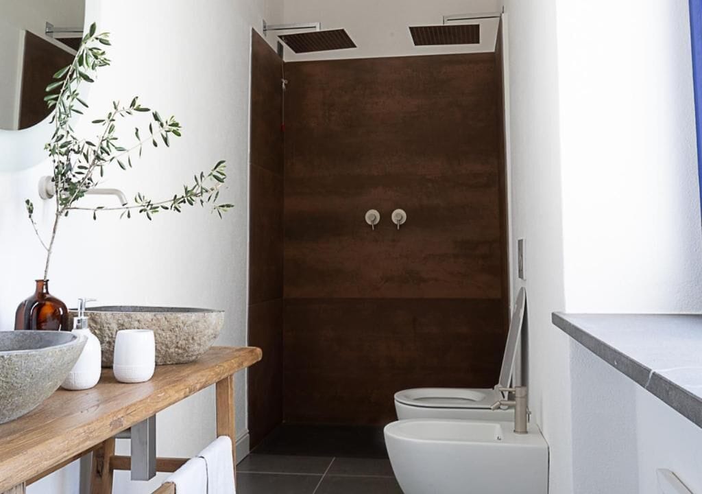 A modern bathroom with a wooden vanity, a large stone basin, a white toilet, and a dark wooden shower enclosure.