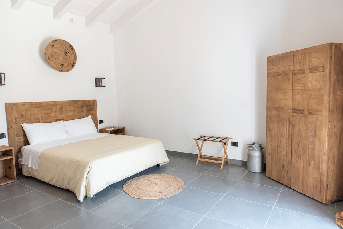 A bedroom with a large bed, a wooden wardrobe, a round woven rug, and a metal container.