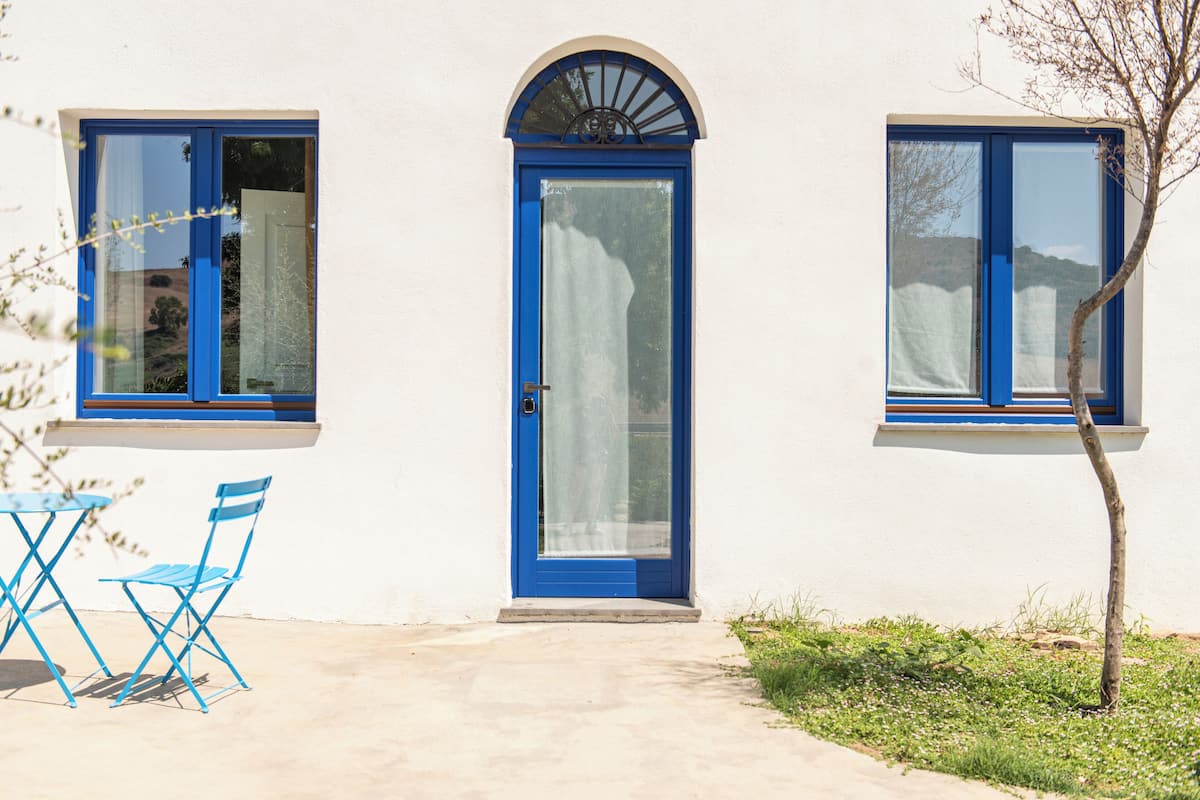A building with a blue door and windows, a small outdoor seating area with blue chairs, and a tree in front.