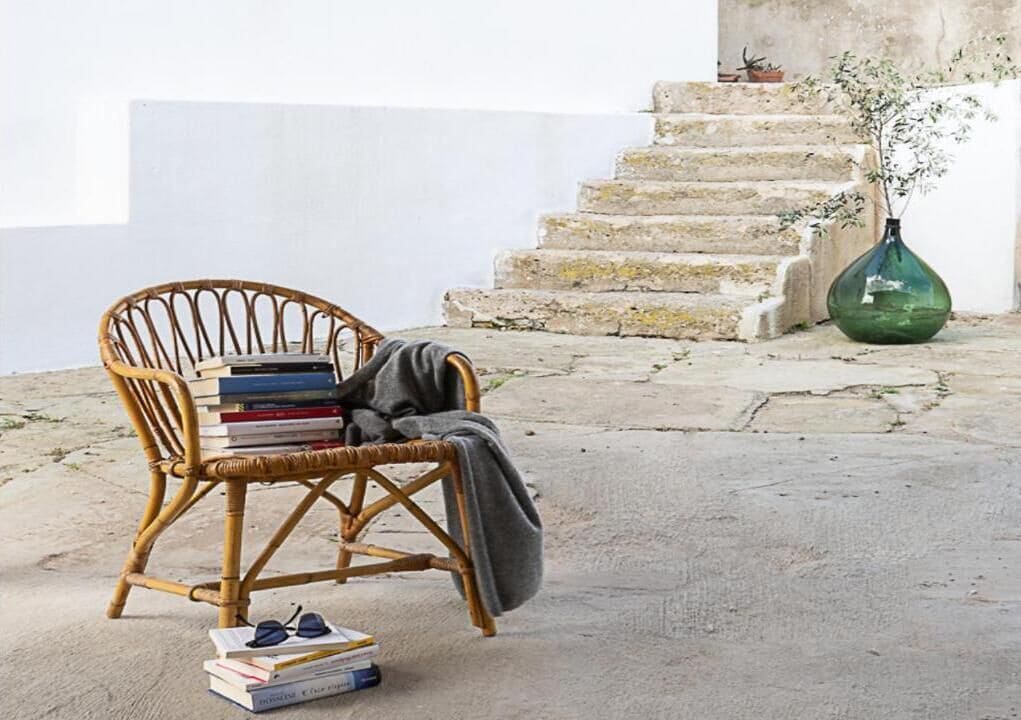 A wicker chair with a stack of books and a pair of glasses on the ground, next to a green vase on a concrete surface.
