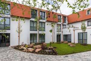 Un edificio moderno con un patio que incluye un sendero pavimentado, una zona ajardinada y varios árboles jóvenes.