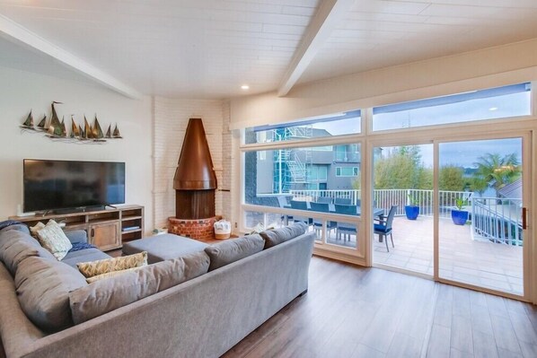 A living room with a large sofa, a flat-screen TV, a brick fireplace, and a balcony with outdoor furniture.