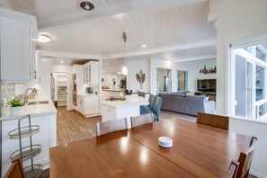 A modern kitchen with white cabinetry, a dining area with a wooden table, and a living room with a grey sofa.