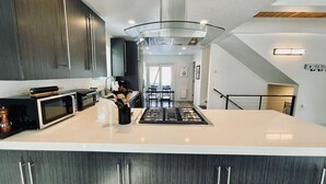 A modern kitchen with dark wood cabinets, a large island, a stainless steel range hood, and a dining area with a table and chairs.