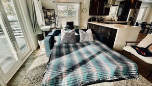 A modern bedroom with a large bed, a small desk, and a kitchenette in the background.