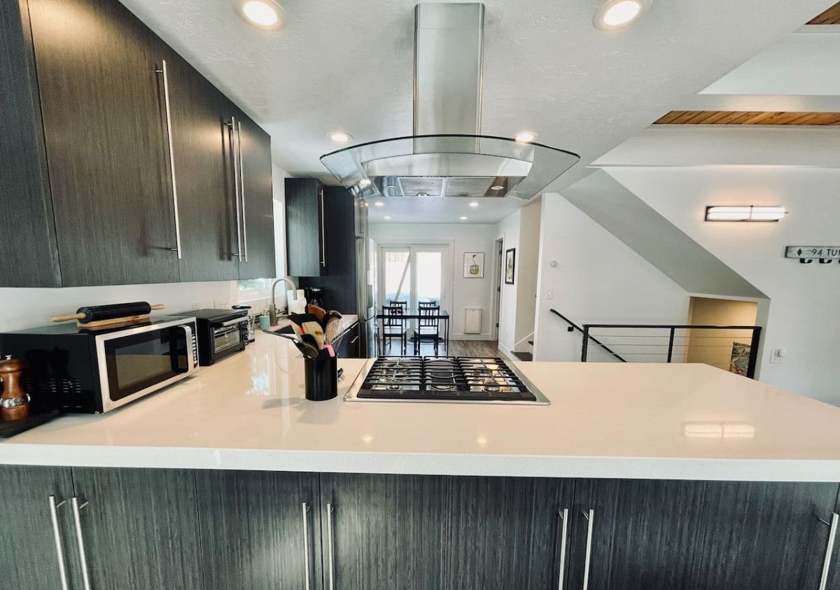 A modern kitchen with dark wood cabinets, a large island, a stainless steel range hood, and a dining area with a table and chairs.