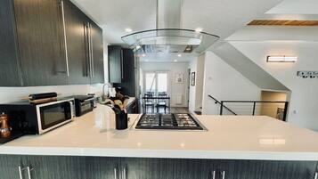 A modern kitchen with dark wood cabinets, a large island, a stainless steel range hood, and a dining area with a table and chairs.