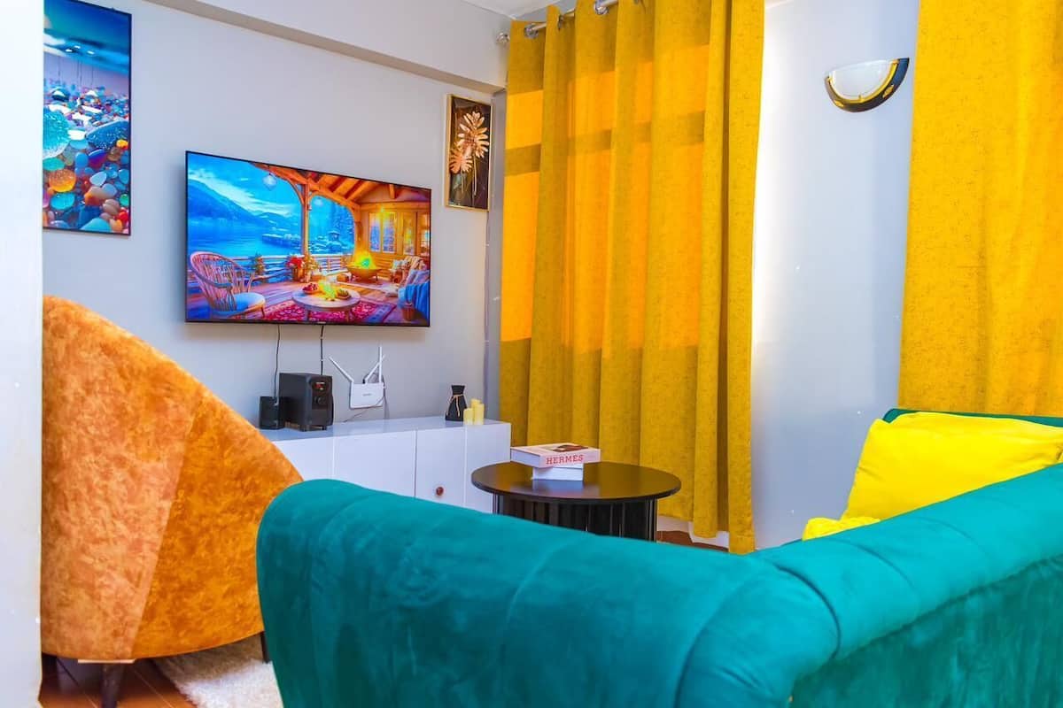 Ein Wohnzimmer mit Fernseher, gelben Vorhängen, einer Couch und einem Tisch mit einer Box Hermes Pralinen.
