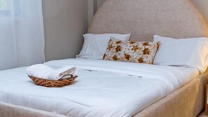 A neatly made bed with a headboard, white bedding, and a basket of folded towels. - CH Apartments Kisii (Kisii)