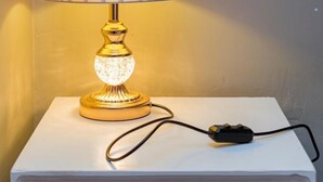 A table lamp with a pleated lampshade and a crystal base, placed on a white nightstand with a power cord. - CH Apartments Kisii (Kisii)