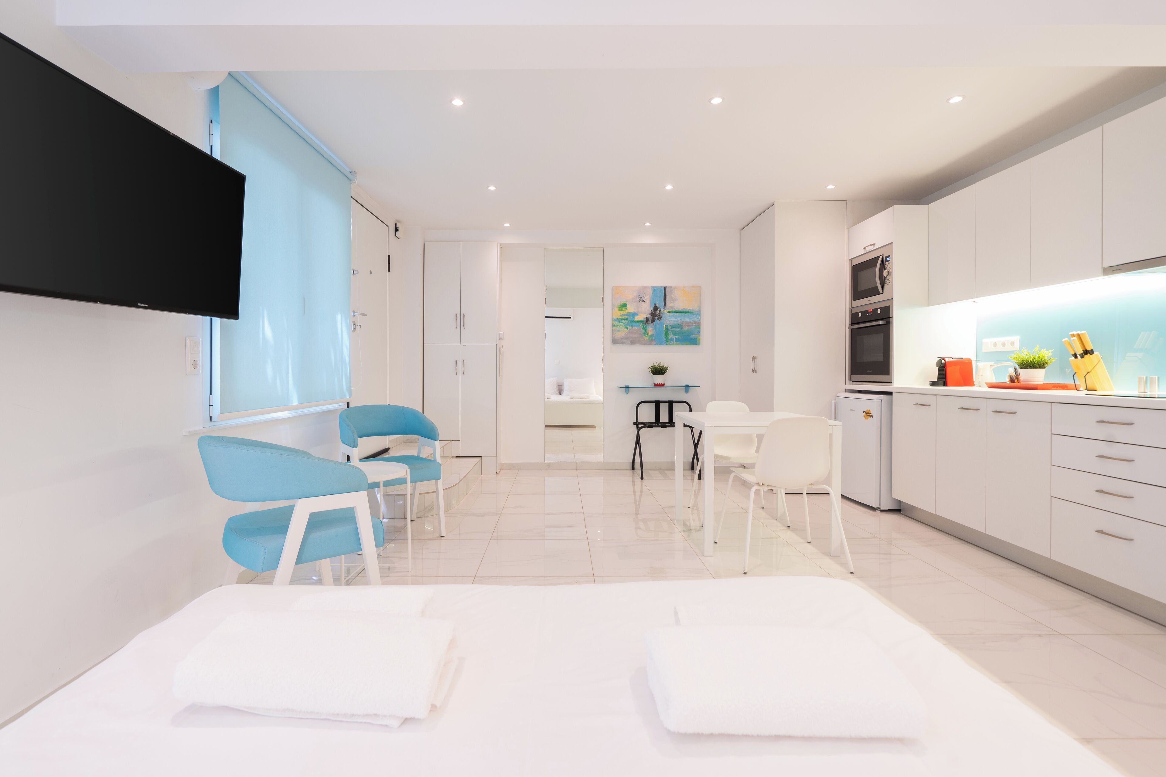 A modern hotel room with a large bed, a desk, a chair, a TV, a kitchen area, and a bathroom.