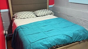 A bed with a teal bedspread and patterned pillows, a wooden headboard, and a window with patterned glass.