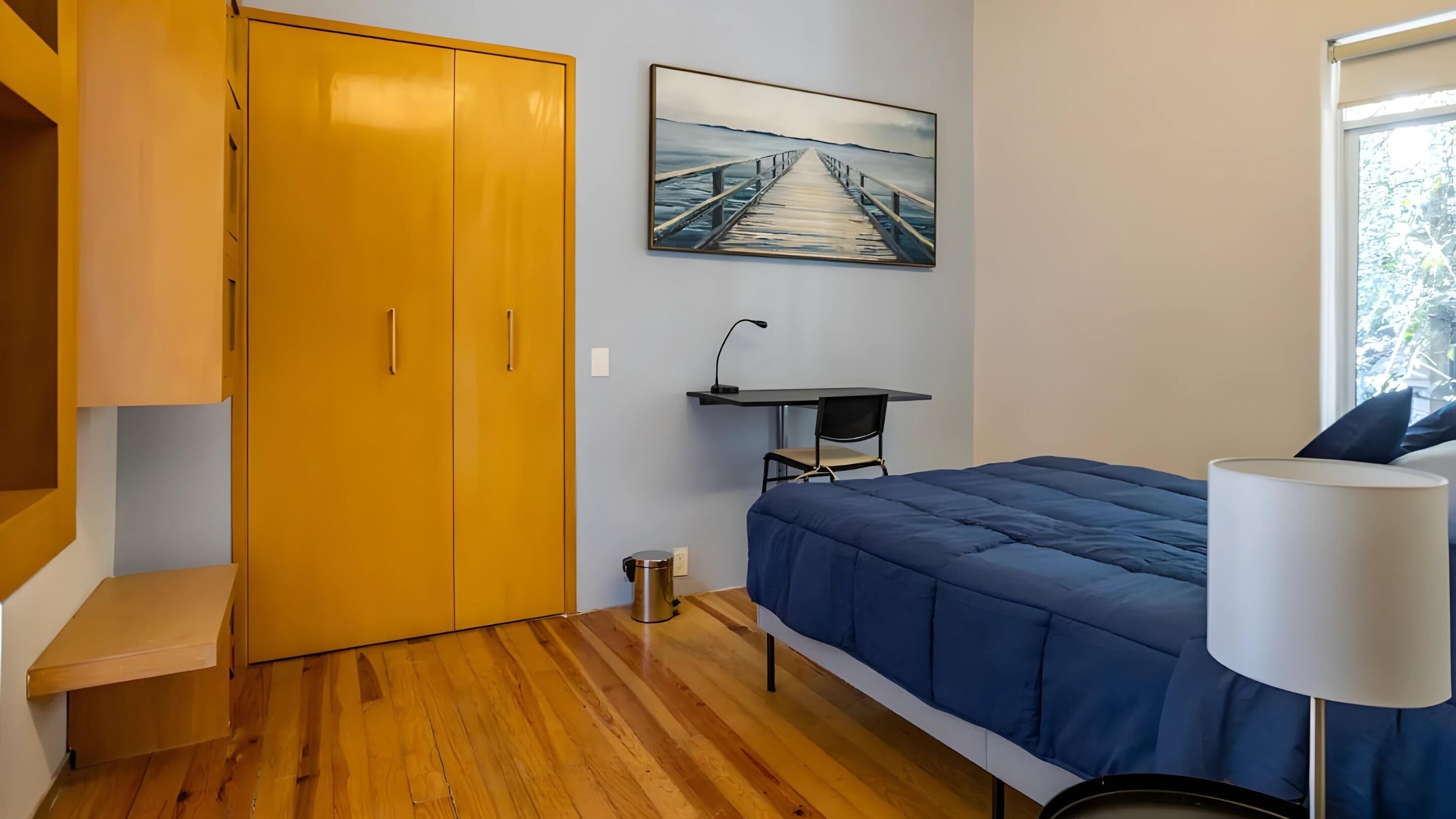 A bedroom with a wooden floor, a bed with a blue duvet, a desk with a chair, a painting of a pier, and a wardrobe.