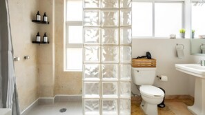 A bathroom with a glass partition, a toilet, a sink, and shelves with toiletries.