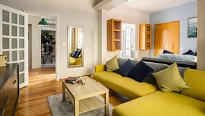 A modern living room with a yellow sofa, a wooden coffee table, and a dining area with a small table and chairs. There is a large mirror on the wall, a wooden cabinet, and a bed with a blue headboard in the background. - Cozy Condesa (Mexico City)