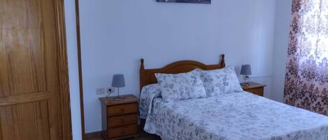 A bedroom with a wooden bed, a nightstand with a lamp, a wall-mounted picture, and a door.