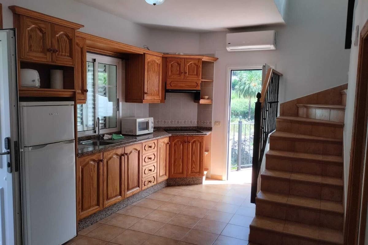 A kitchen with wooden cabinets, a refrigerator, a microwave, and a window overlooking a garden.