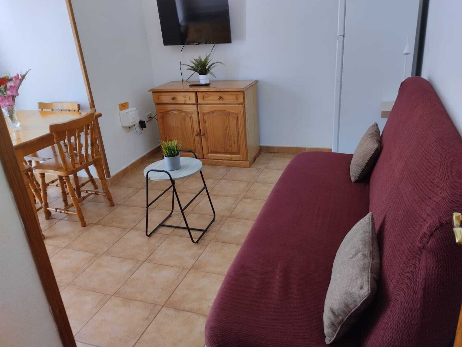 A small living area with a sofa, a dining table with chairs, a small stool, a cabinet, and a TV mounted on the wall.