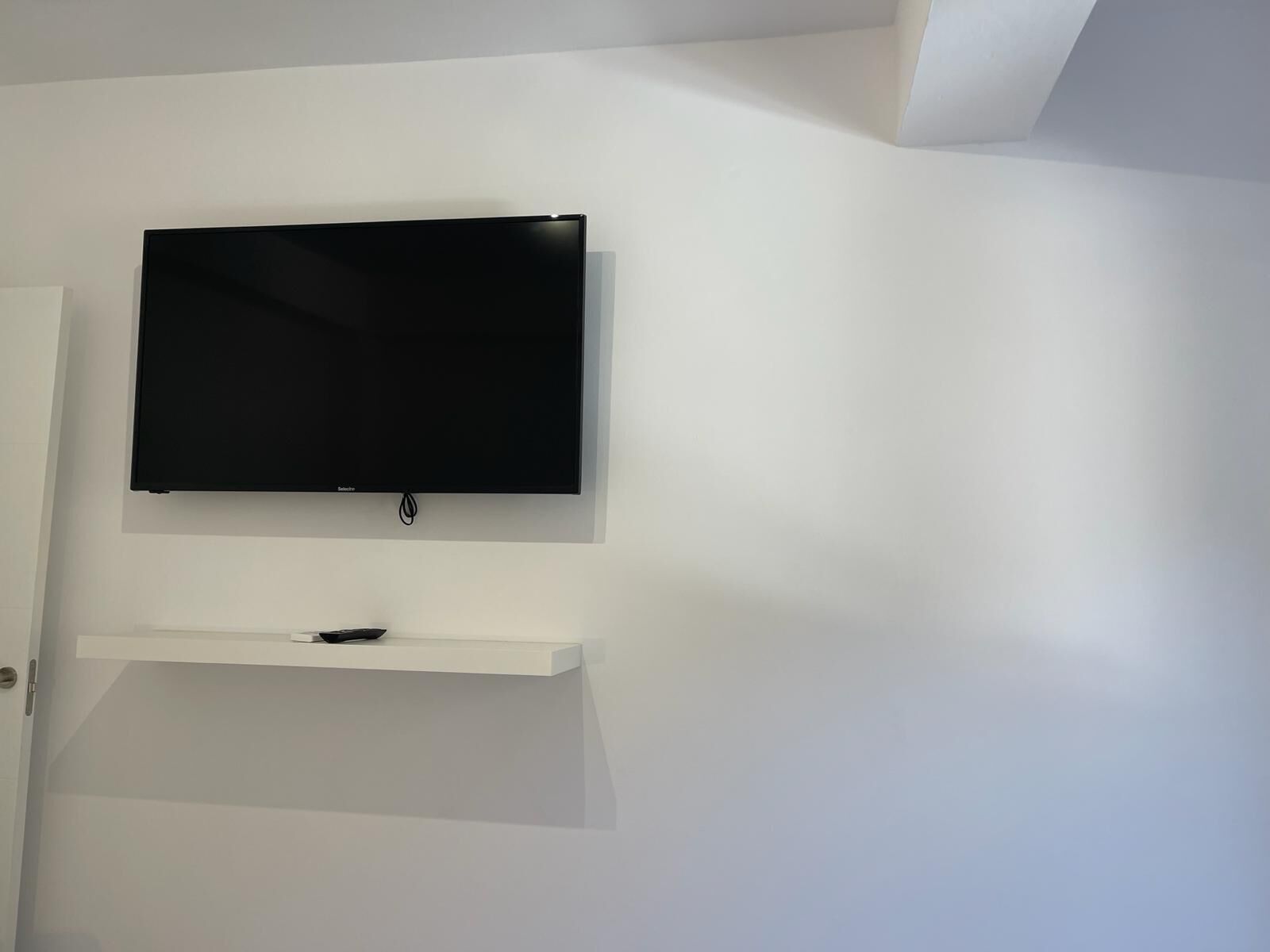 Un televisor de pantalla plana montado en una pared blanca, con una puerta blanca a la izquierda y un estante blanco debajo.