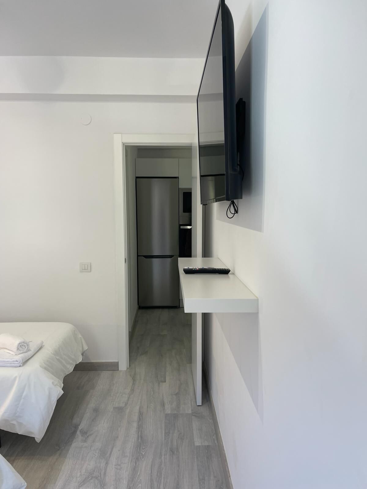 Une chambre d’hôtel moderne équipée d’un lit, d’un téléviseur à écran plat fixé au mur, d’une petite table blanche et d’un réfrigérateur encastré.