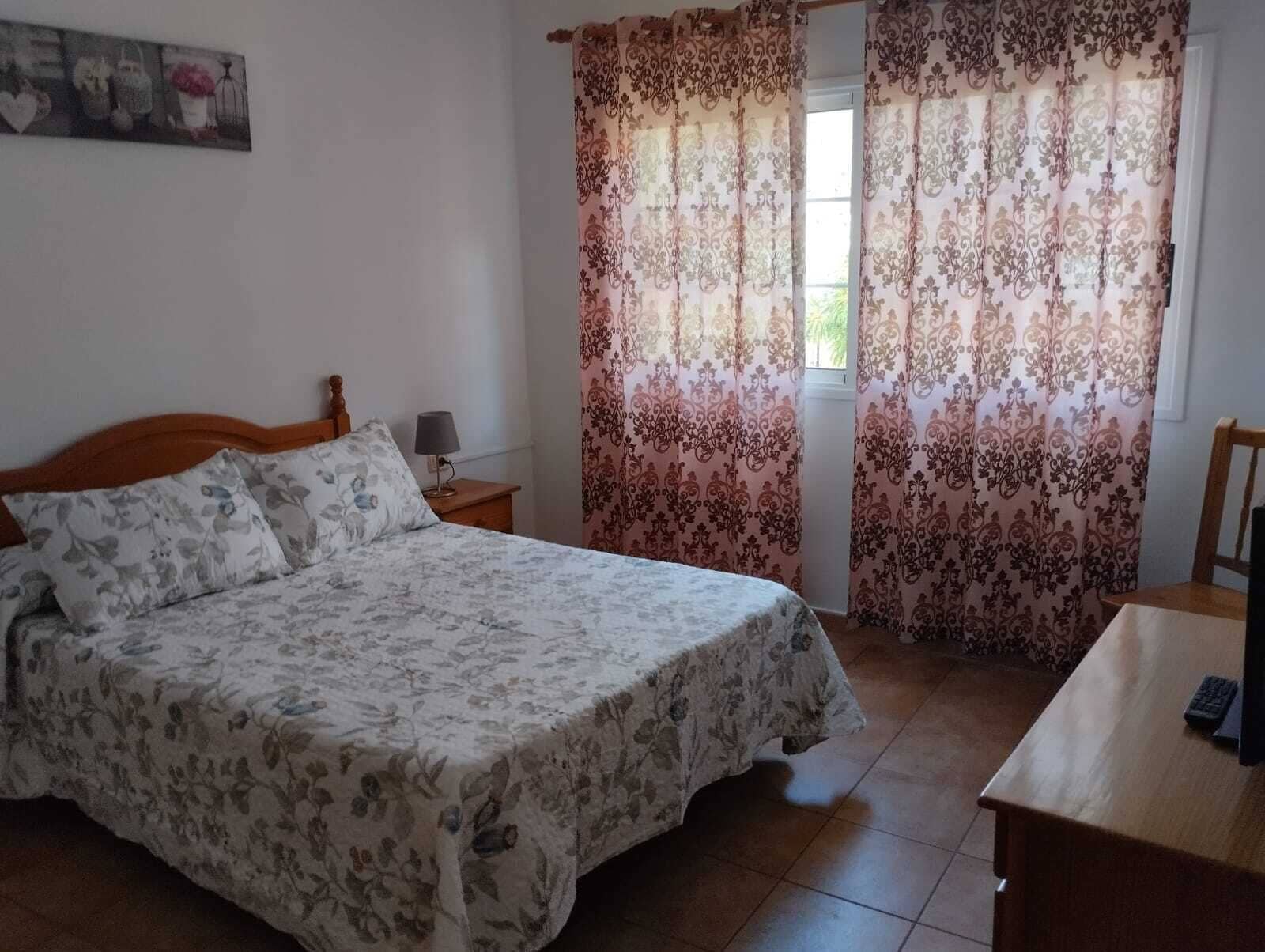 A bedroom with a bed, a nightstand, a desk, and curtains.