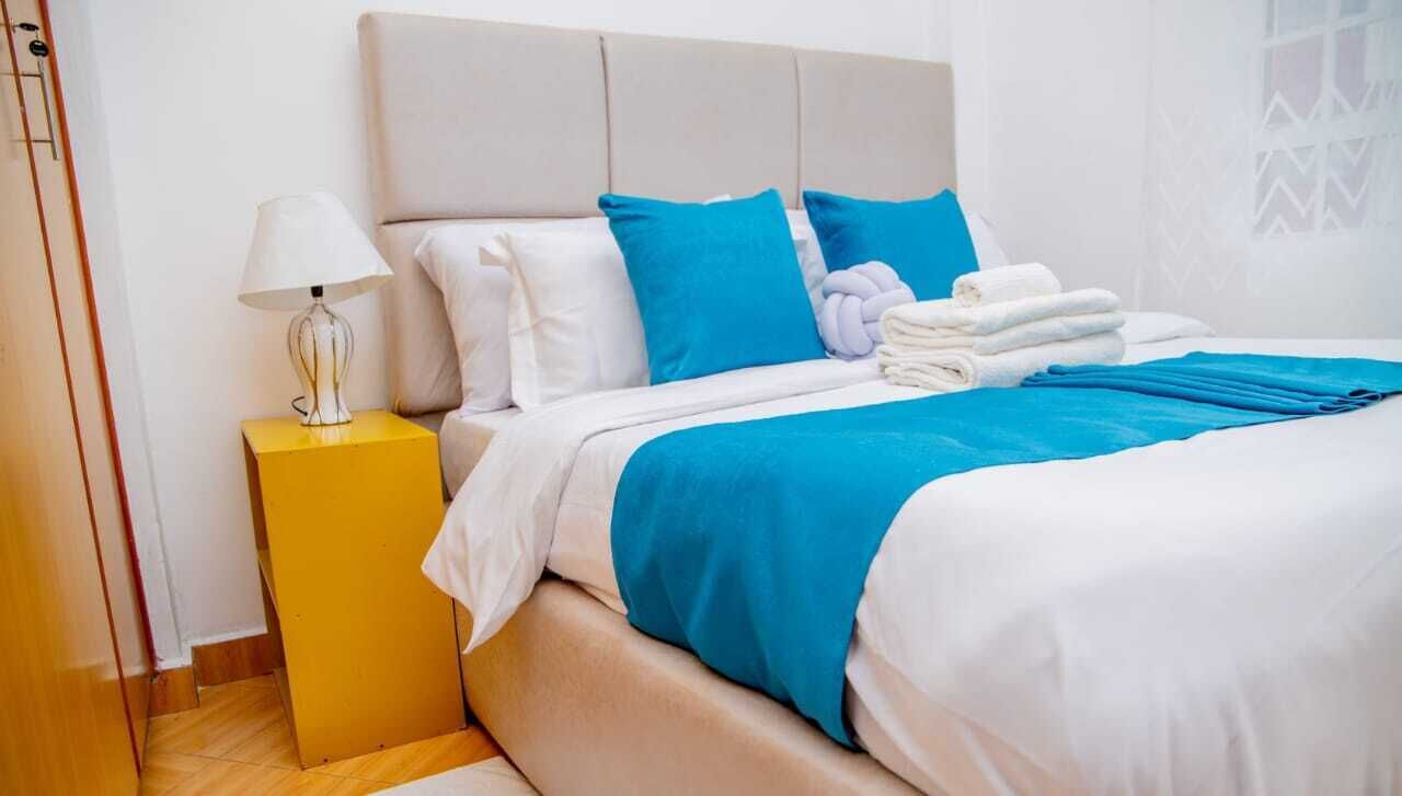 Cama bem arrumada com roupa de cama branca e azul, uma luminária sobre uma mesa de cabeceira amarela e uma toalha dobrada na cama.