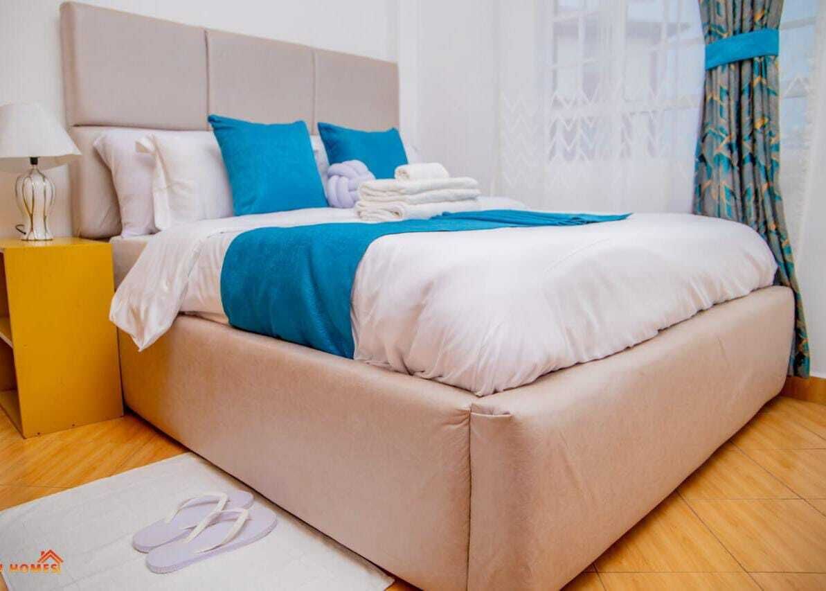 Una cama bien hecha con cabecera color beige, ropa de cama azul y blanca, y un par de toallas blancas a un lado.