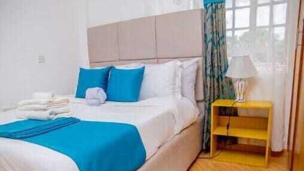 A neatly made bed with blue and white bedding, a bedside table with a lamp, and a window with curtains. - Lux Suites Truna Executive Homes Kisii (Kisii)