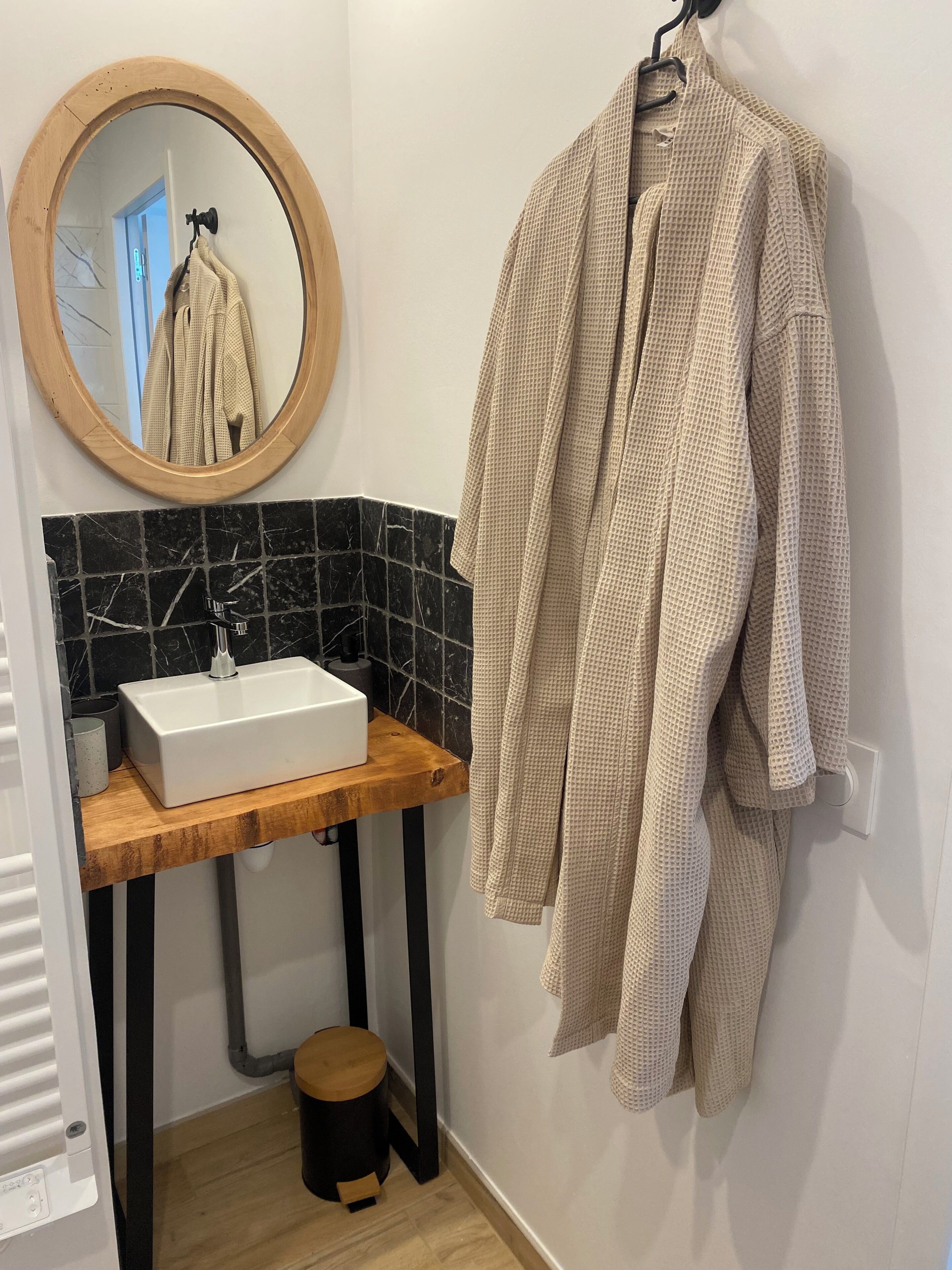 Une salle de bain avec un meuble-lavabo en bois, un lavabo blanc, des carreaux de marbre noir, un miroir rond et une serviette beige accrochée au mur.