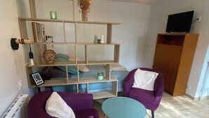 A room with two purple armchairs, a small round table, a wooden shelving unit with various decorative items, and a hanging light fixture.